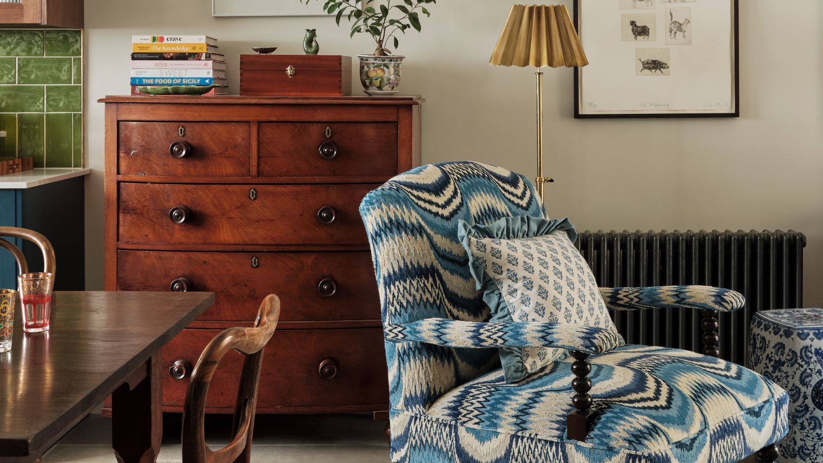 An open plan kitchen diner, with just a corner of a kitchen cabinet in view with green subway splashback tiling. Front and center is a patterned upholstered armchair in blue and cream, with bobbin wood feature. In the background there is a black radiator visible, as well as a mid-century five-drawer cabinet