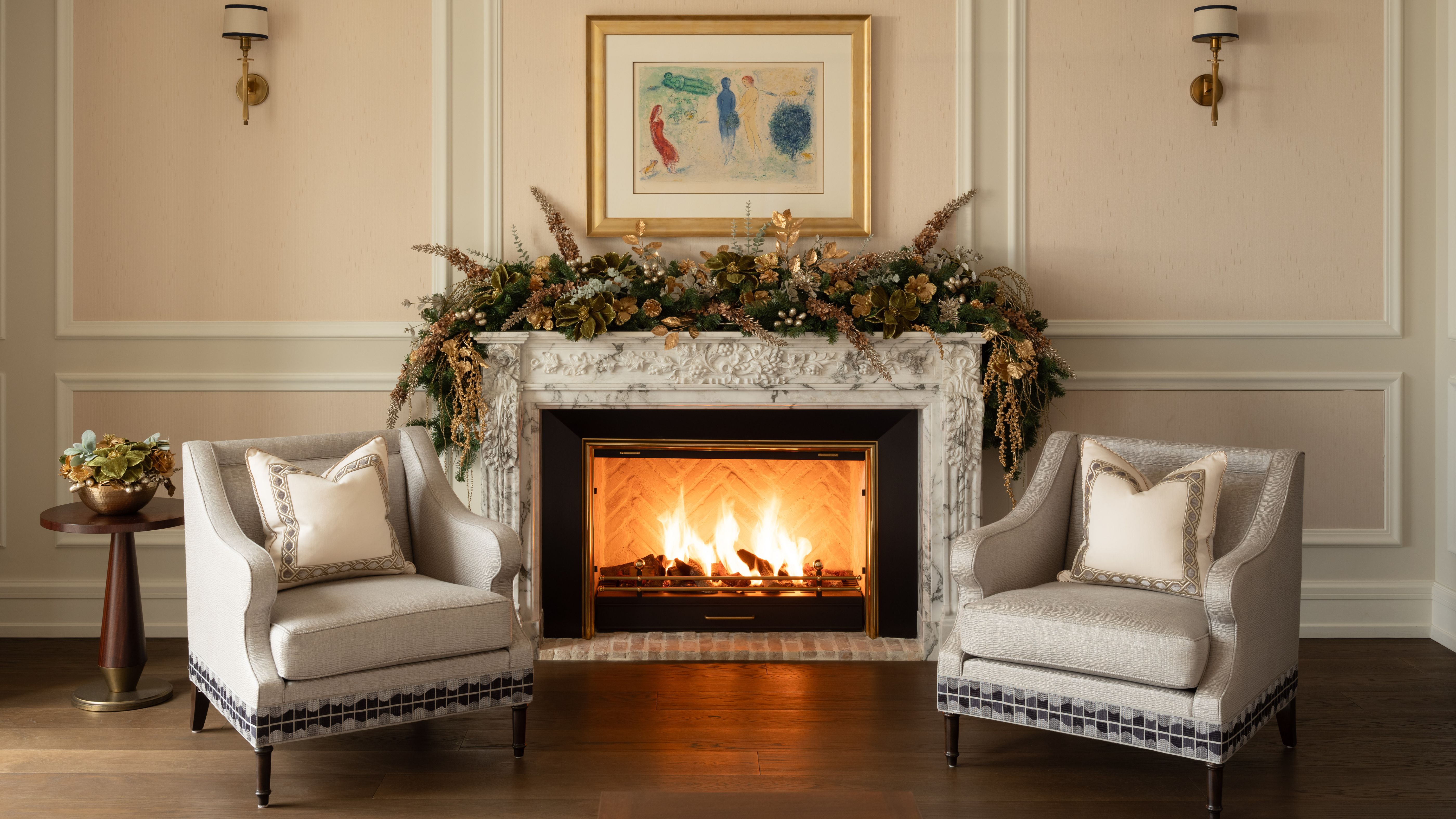 Opulent festive living room featuring ornate picture frame molding, grey accent chairs, lush garland, and a roaring fire