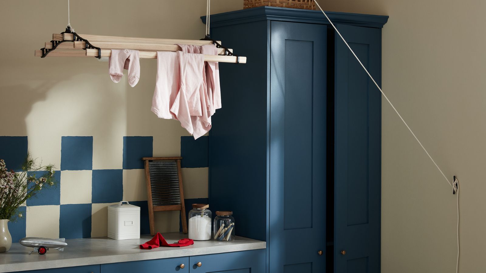 Dark blue painted cabinets with matching blue and white tiles in a small laundry room. A ceiling-suspended drying rack above, with some pink clothes on it. 