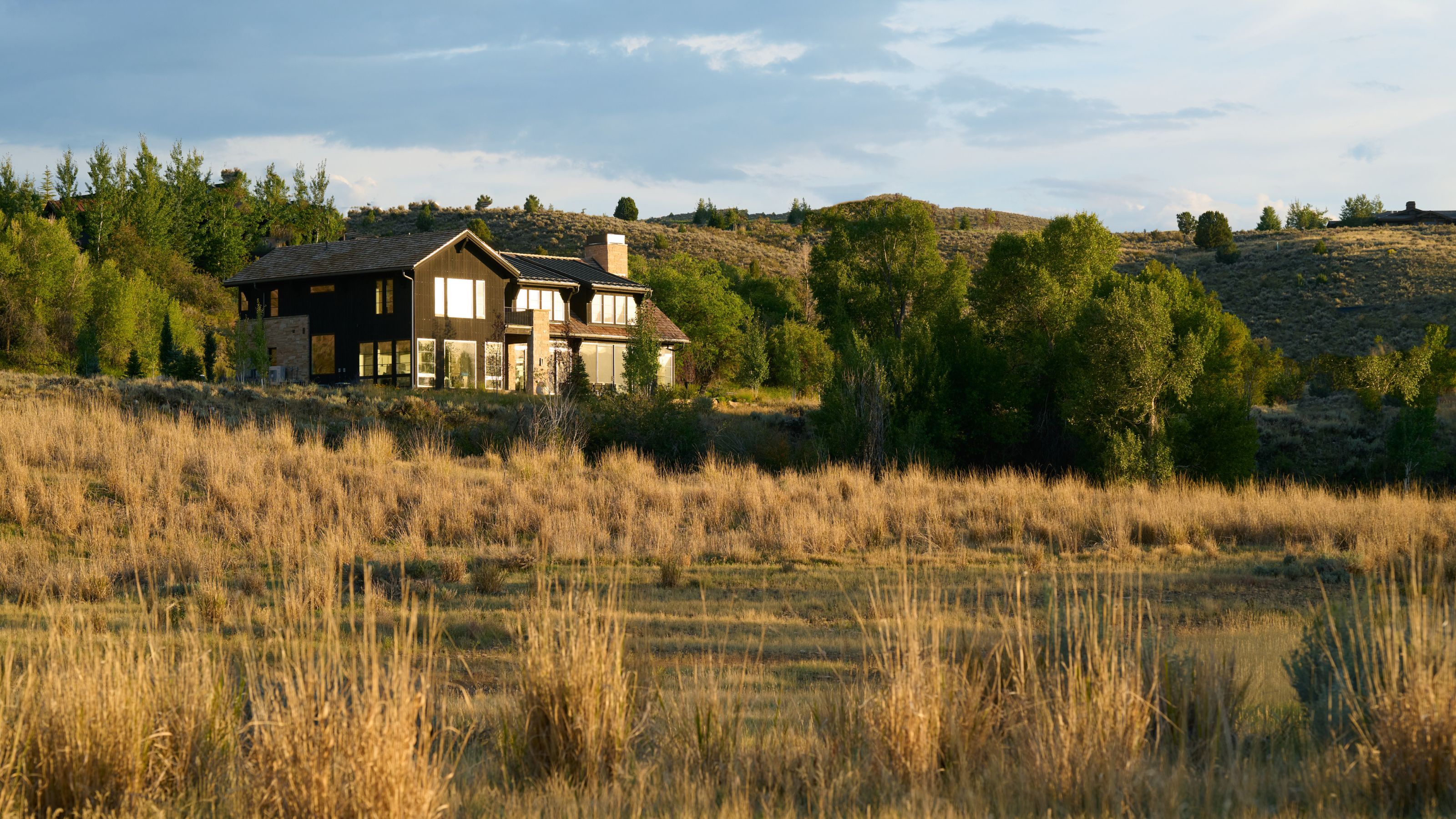 A modern stone and wood house surrounded by tall grasses and trees, set against rolling hills at sunset