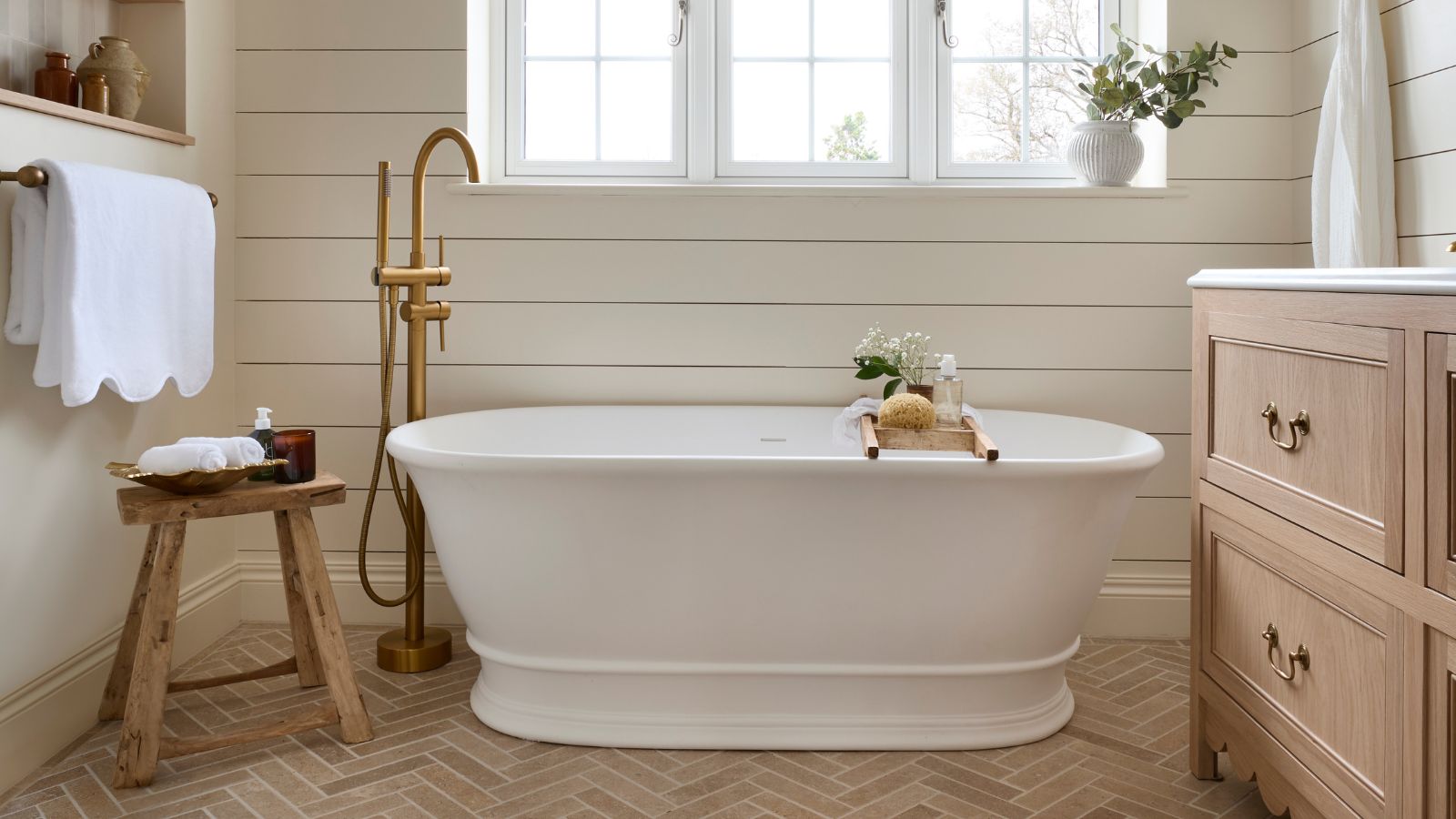 paneled cream bathroom with a light oak scalloped vanity, white freestanding bath with a wooden stool 