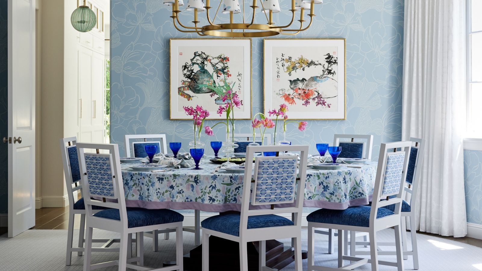 Dining room with floral blue wallpaper and a circular dining table. Blue and white chairs sit around the table and a gold chandelier hangs above