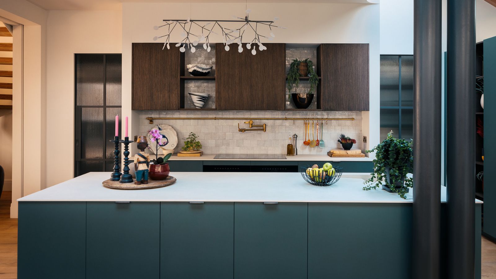 A blue kitchen with large island with white countertop, stove top with white Zellige tiled backsplash with wooden cabinets above, a statement lighting fixture, and two exposed beams to the right.