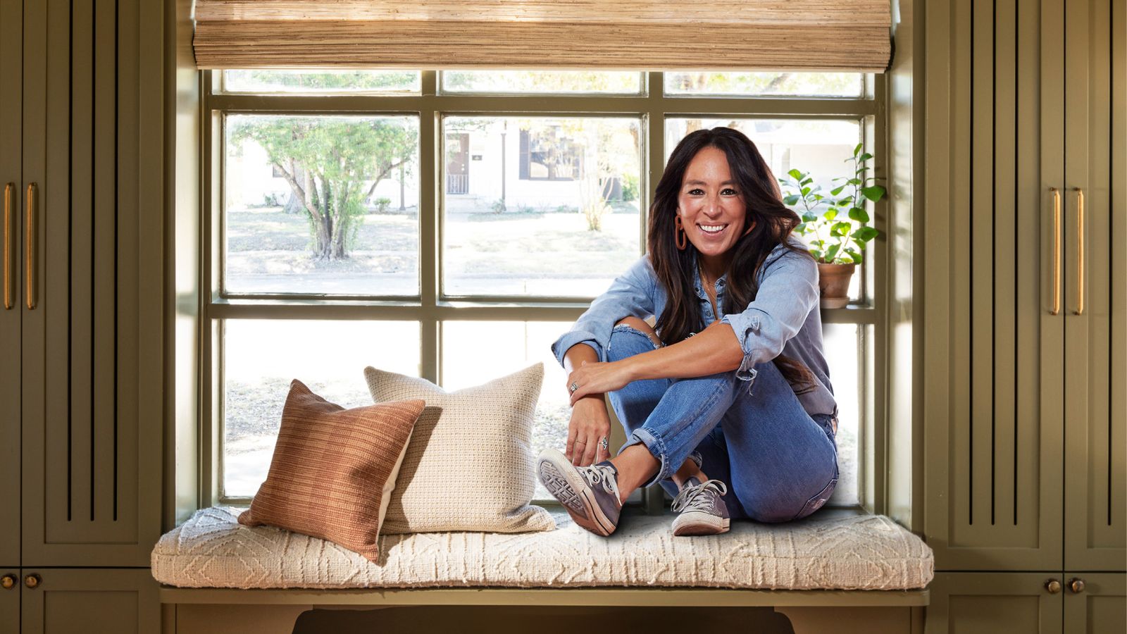 designer Joanna gaines photographed sat on an entryway bench with green cabinetry either side, a bamboo window blind, and two cushions