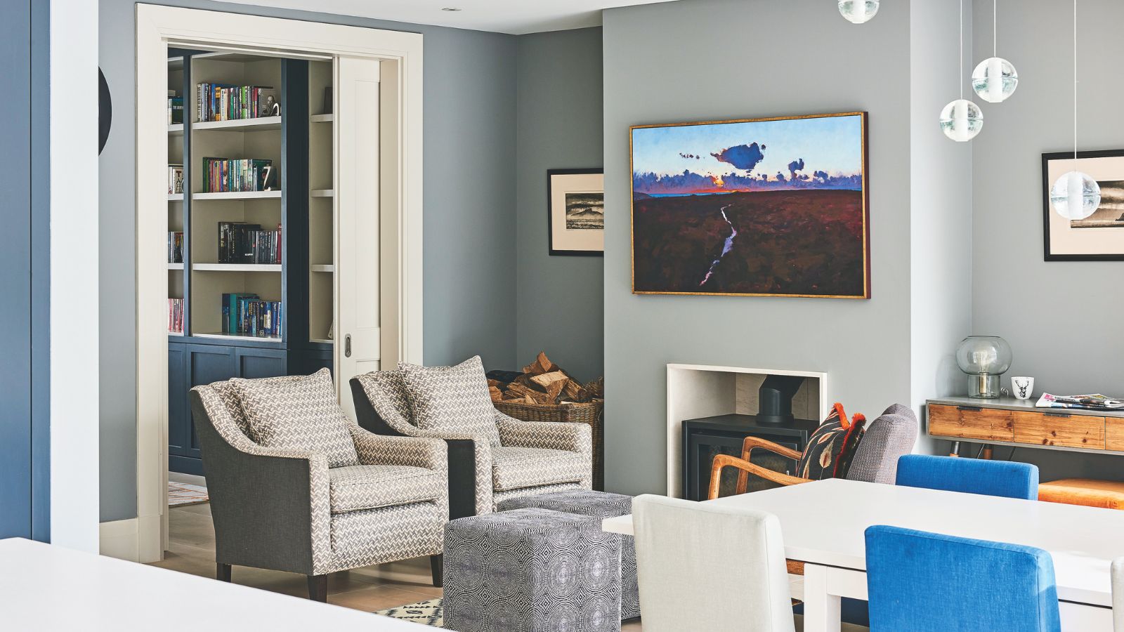 A grey living room space, with a central black log burner with a large artwork above, two matching patterned upholstered chairs, two matching cubic ottomans, and a grey upholstered chair with wooden handles and a patterned cushion. To the right foreground is a white dining table with blue and white chairs. To the left, a room is seen behind the matching upholstered chairs, with blue and white bookshelves.