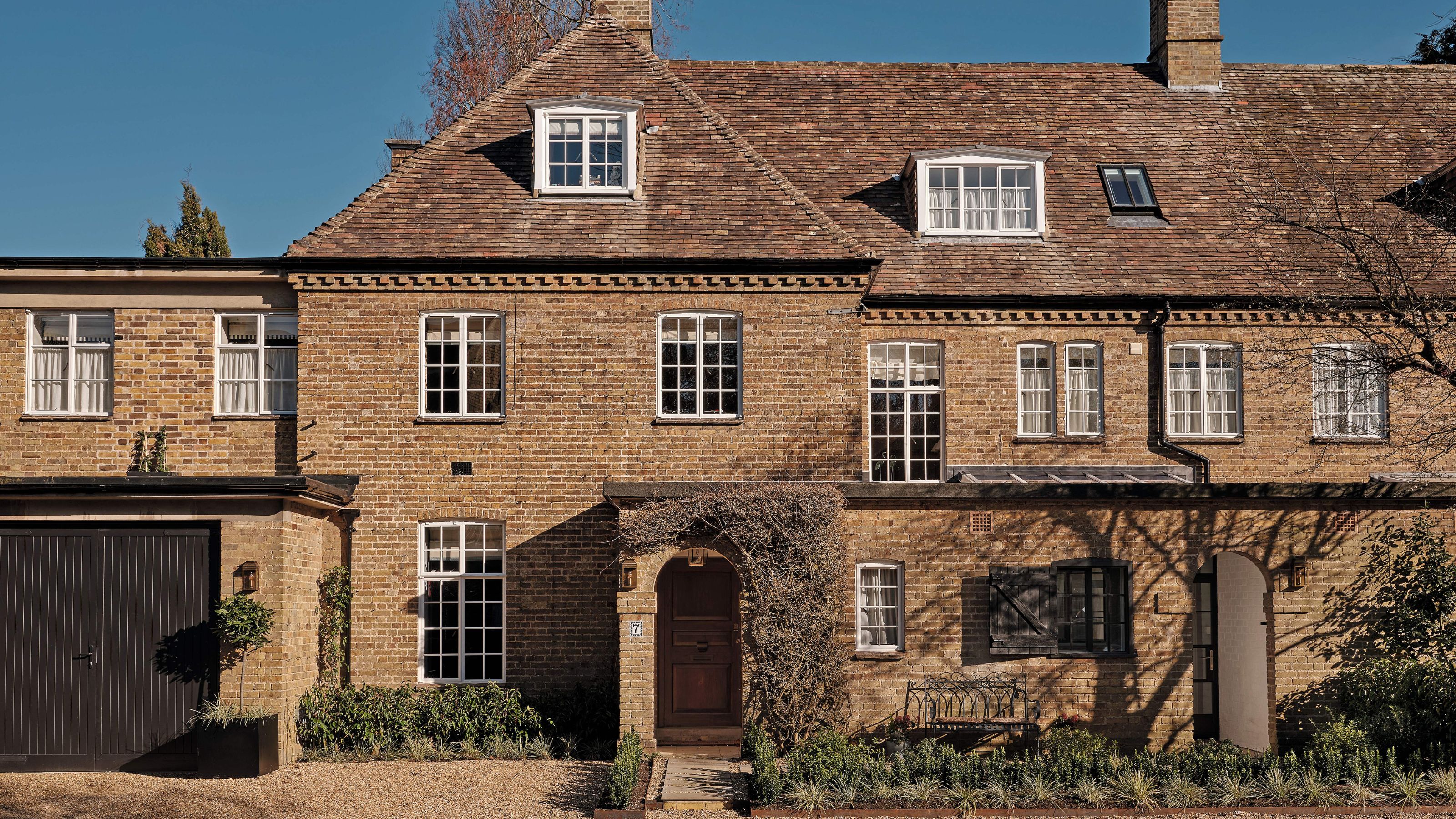 Front view of a traditional brick townhouse with white-framed windows, pitched tile roof, and a wooden front door surrounded by creeping vines