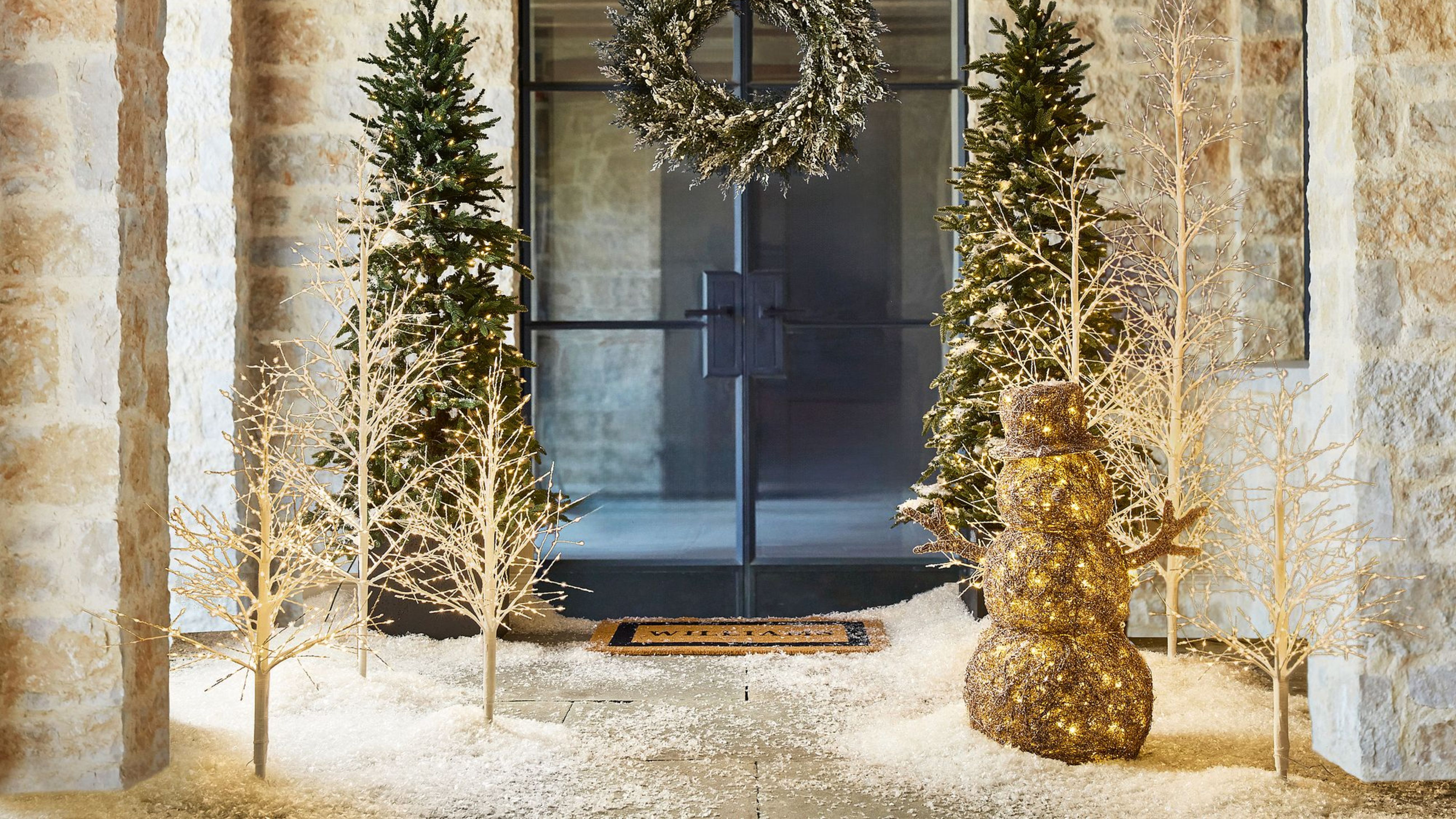 Twinkling twig Christmas trees clustered among traditional evergreens in front of a snow-covered glass entryway, which is punctuated with a coordinating wreath.