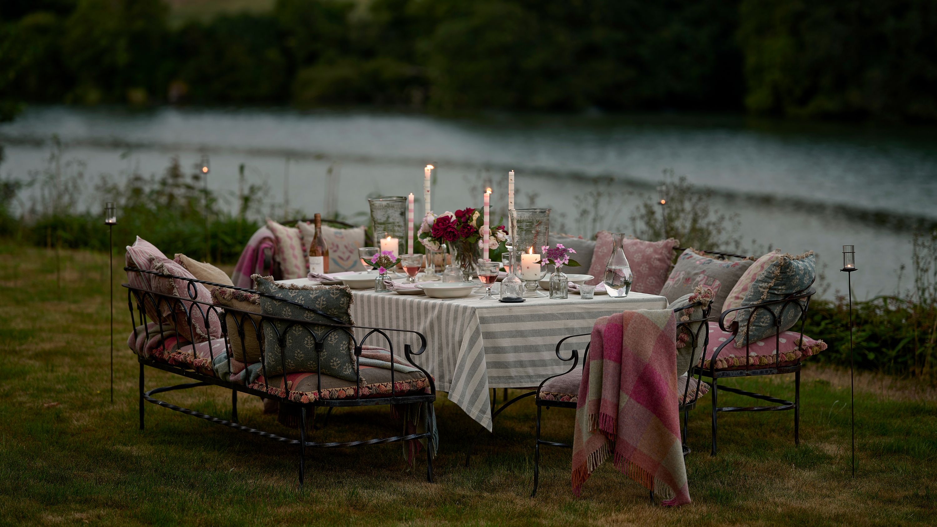 outdoor countryside garden party in the evening shot at dusk by a lake with tall candles lit, wrought iron seating with cushions and blankets