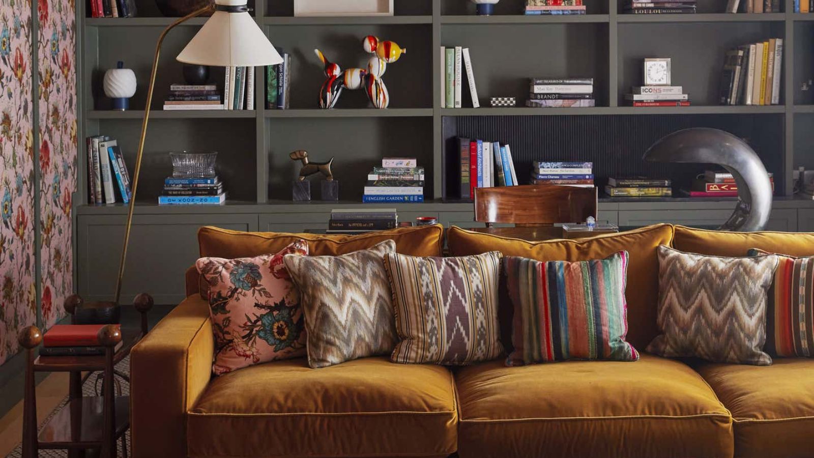A maximalist living room with a mustard velvet sofa in front of built-in grey book shelves. 