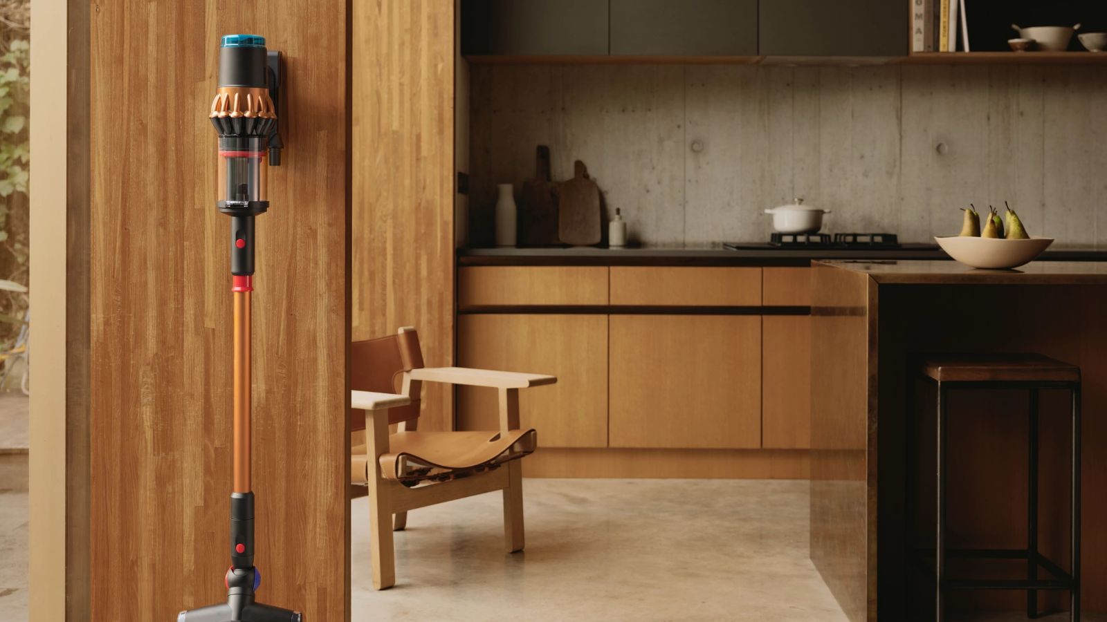 Dyson V16 Piston Animal mounted on wall in modern, wooden kitchen. A kitchen island, wooden armchair, kitchen cabinets, and a countertop with miscellaneous kitchen items are pictured in background.