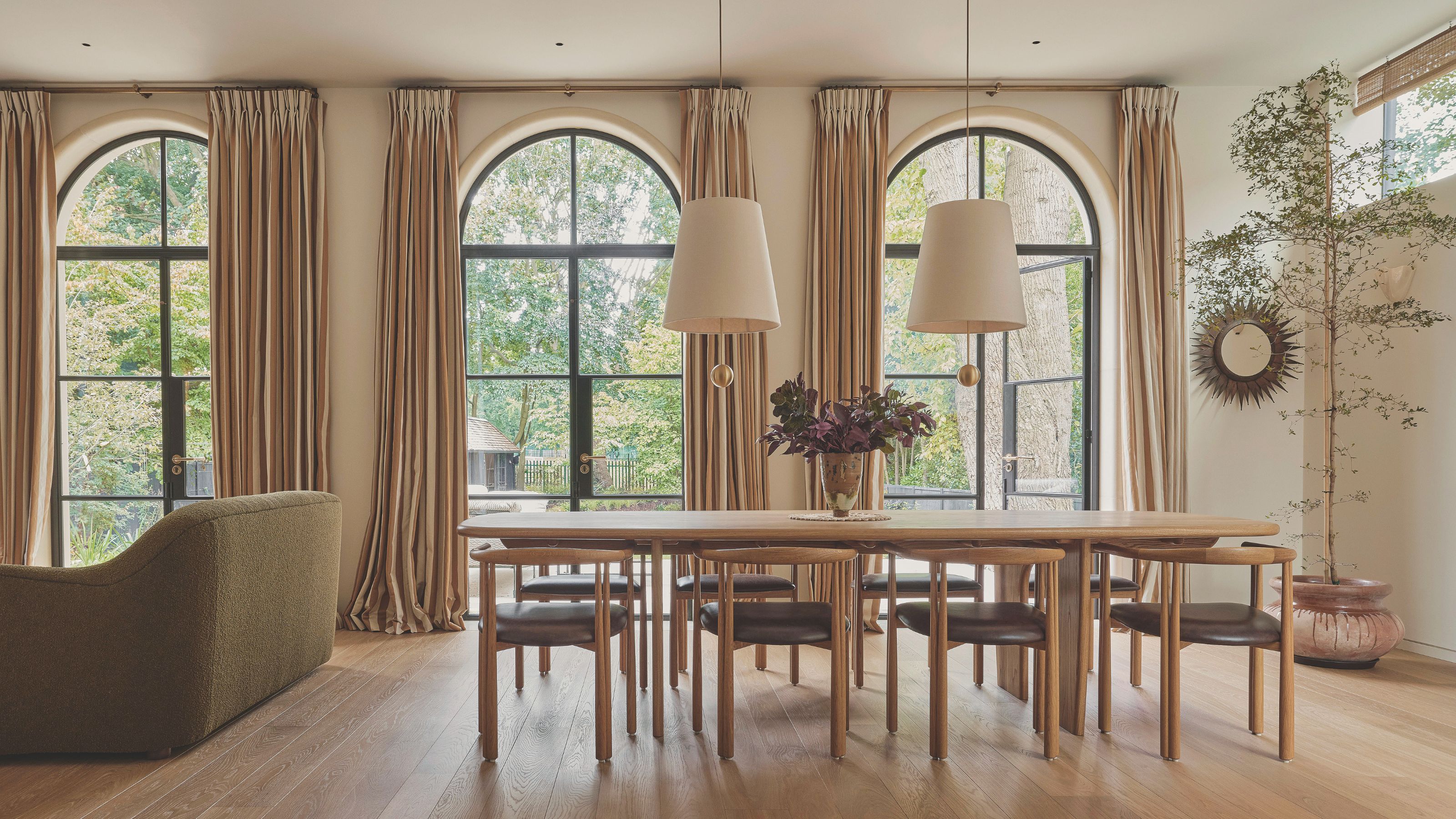 Open plan dining room with three large arched windows each with long cream drapes. A large wooden dining table sits in the middle of the space with two pendant lights with white shades hanging above