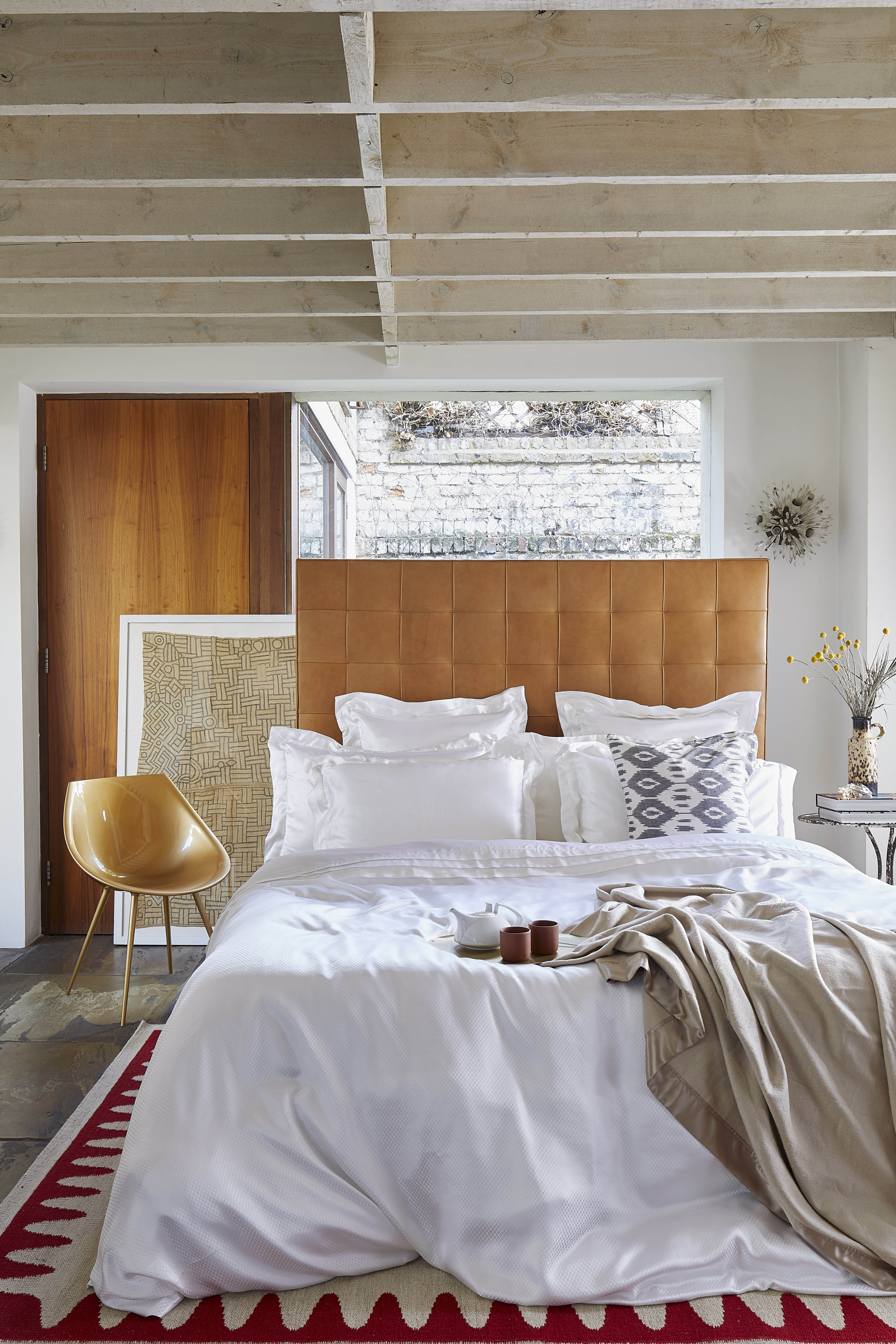 bedroom with beamed ceiling, stone floor, leather headboard, designer modern chair, white silk bedding, white walls 