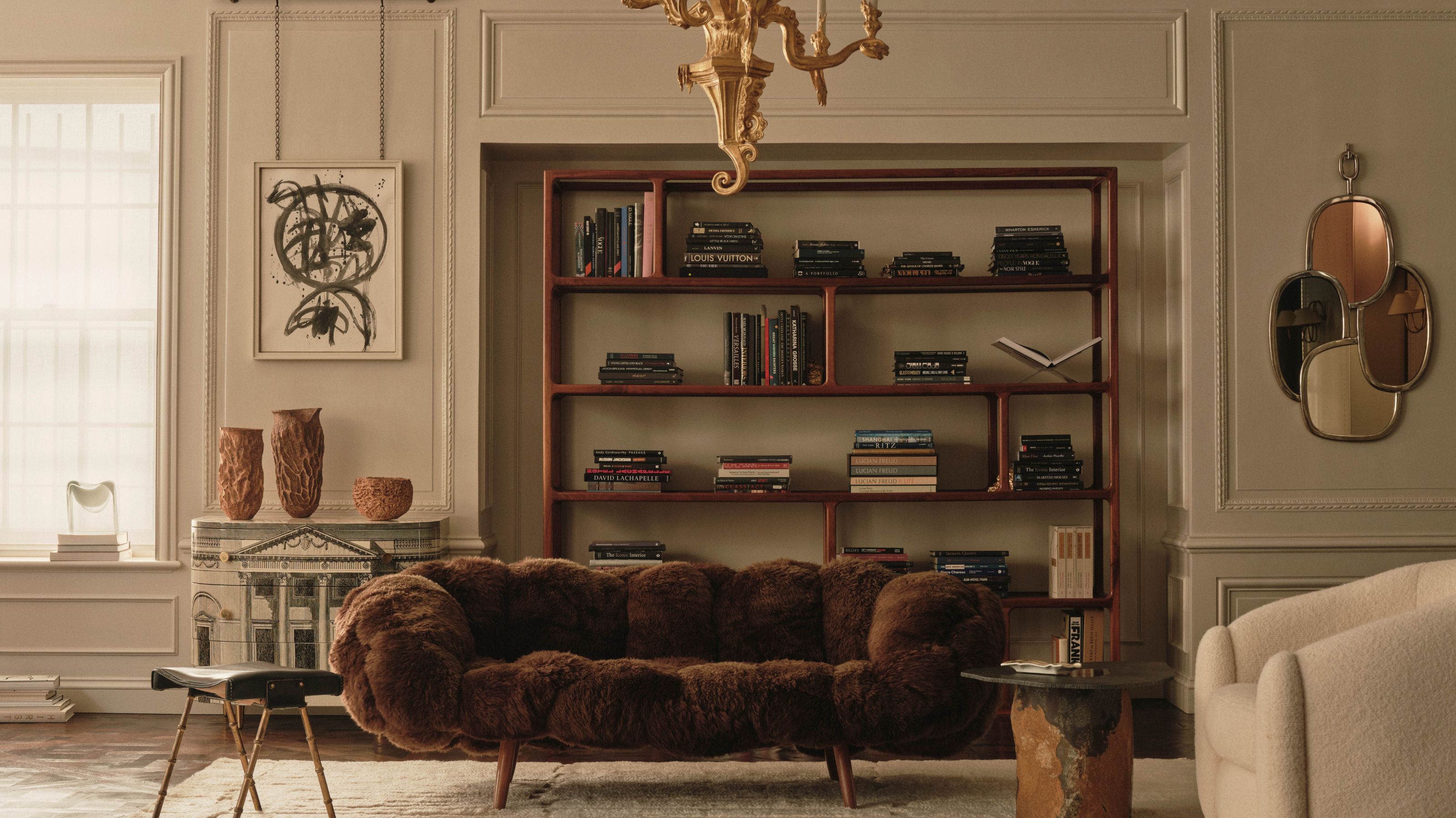 Cream living room with panelled walls and modern furniture, including a brown furry couch, a wooden minimalist bookcase and a cream boucle arm chair