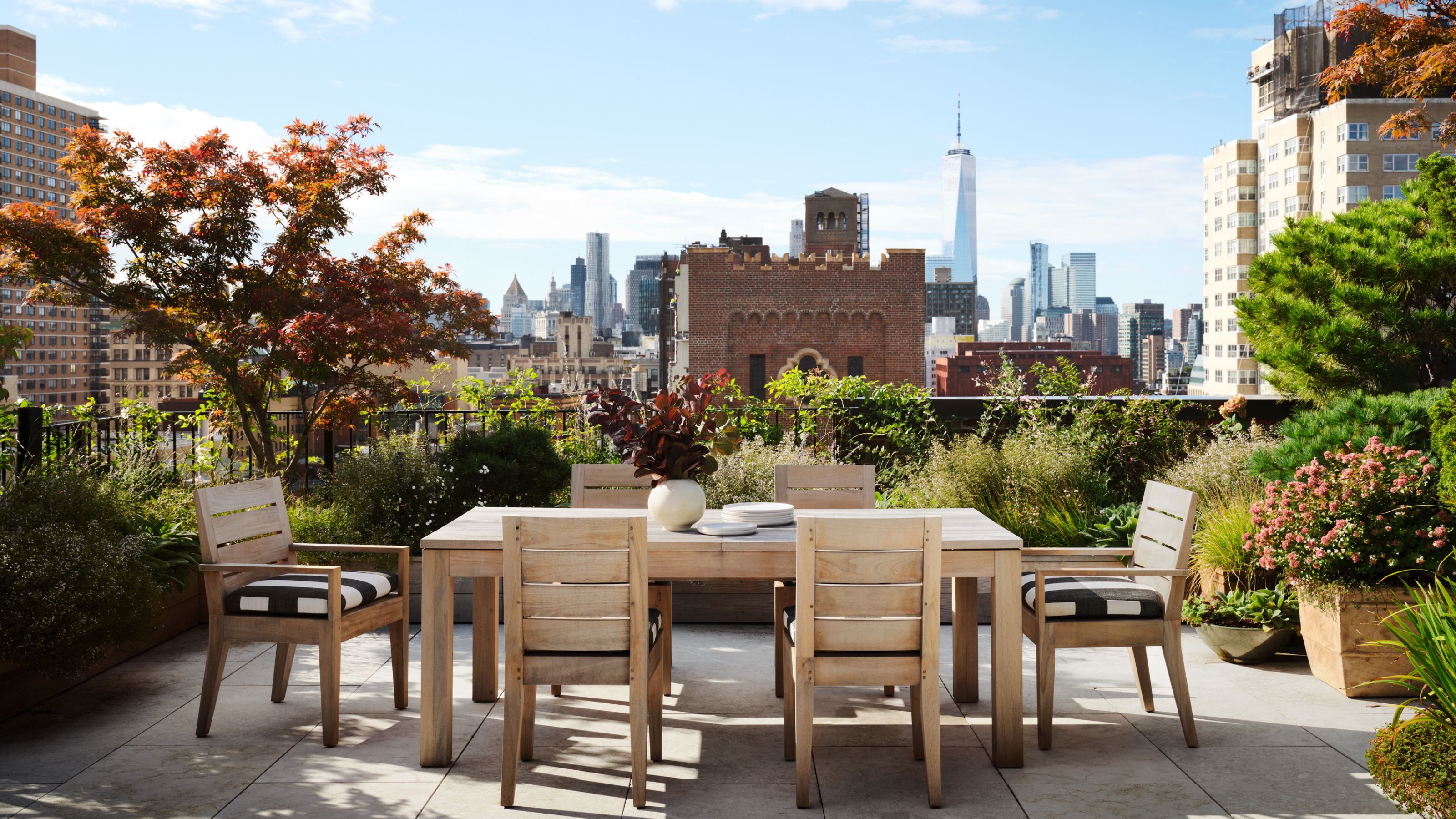 A roof top garden with view of the New York skyline. A wooden table and chairs sits in the middle with mature planting surrounding 