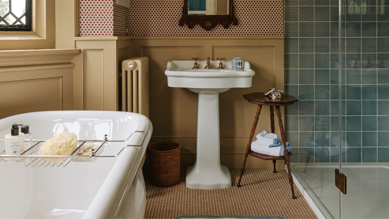 Bathroom with butter yellow panelling, pedestal sink, blue tiled shower, wooden side table and bath with rack 