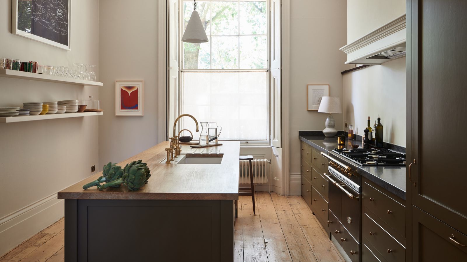 Compact cream galley kitchen with black wooden cabinets and wood-top island and floating wall shelves. There is a large window at the end and a vintage radiator just in view
