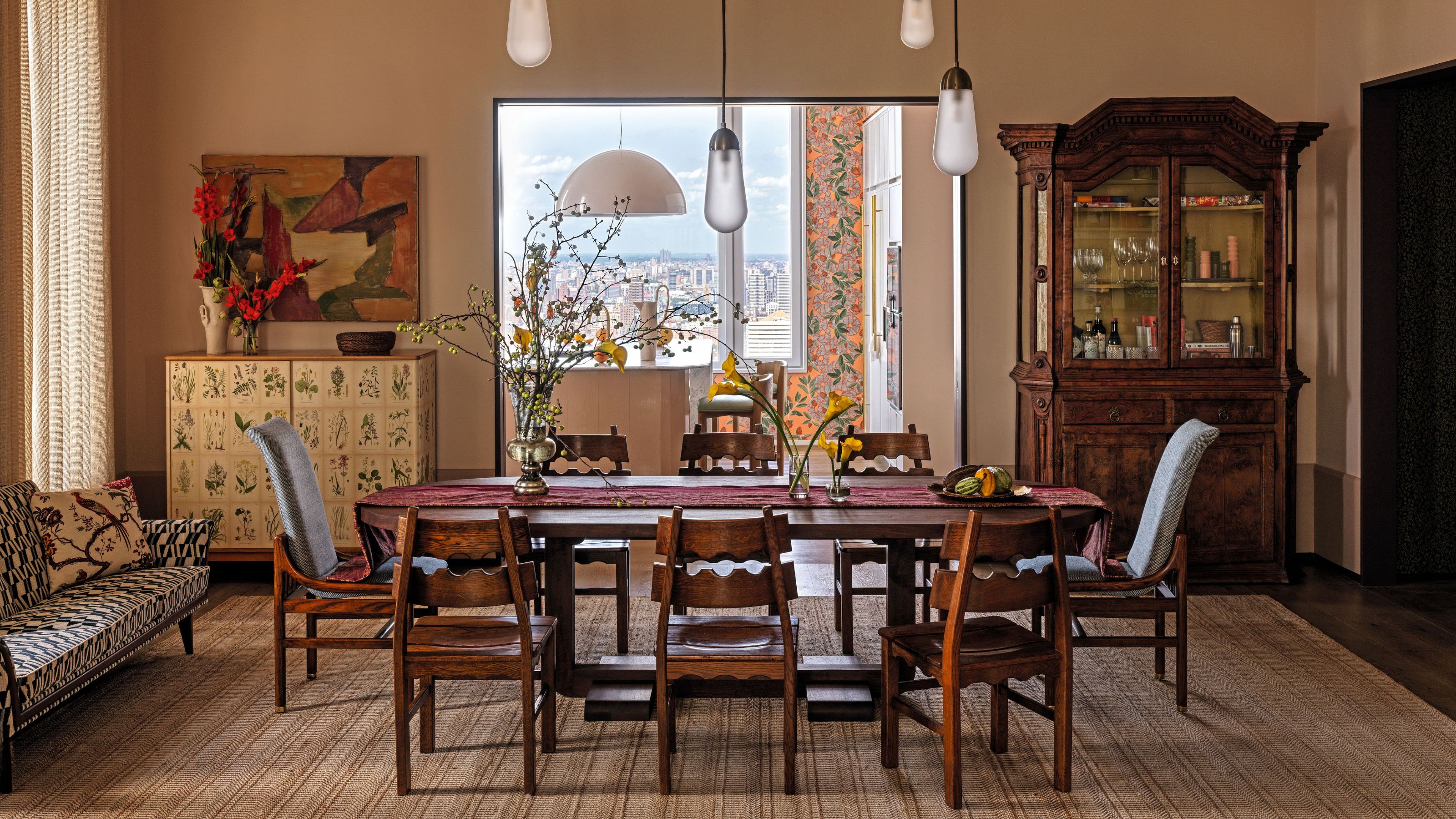 Dining room with creamy plaster pink wallas and a large wooden dining table and vintage wooden chairs in the middle. An antique glass fronted cabinet is in one corner and a vintage sofa under the window