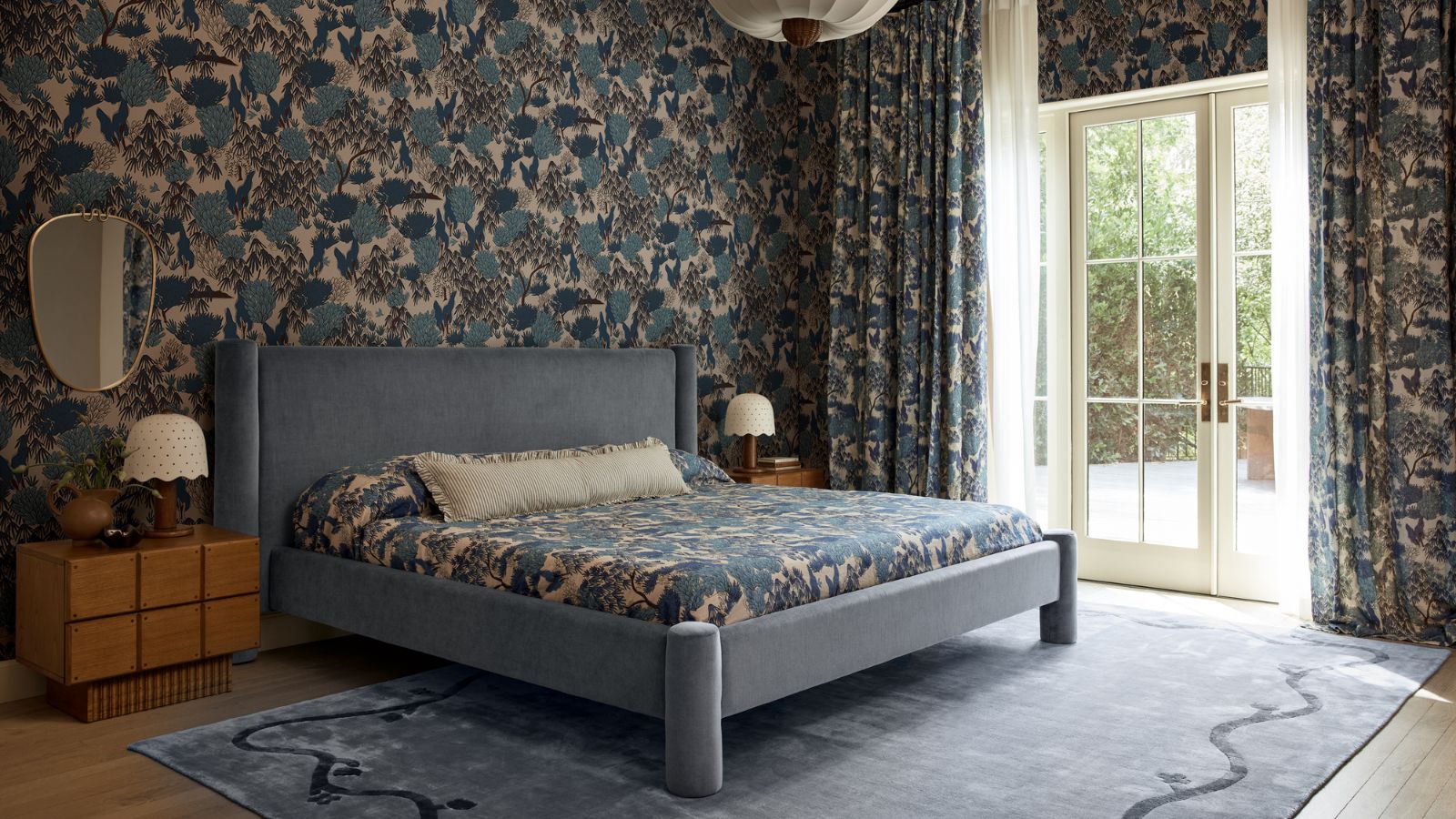 blue pattern drenched bedroom with matching blue floral printed wallpaper, drapes and bed linens with a large blue rug, blue velvet upholstered bed and wooden nightstands