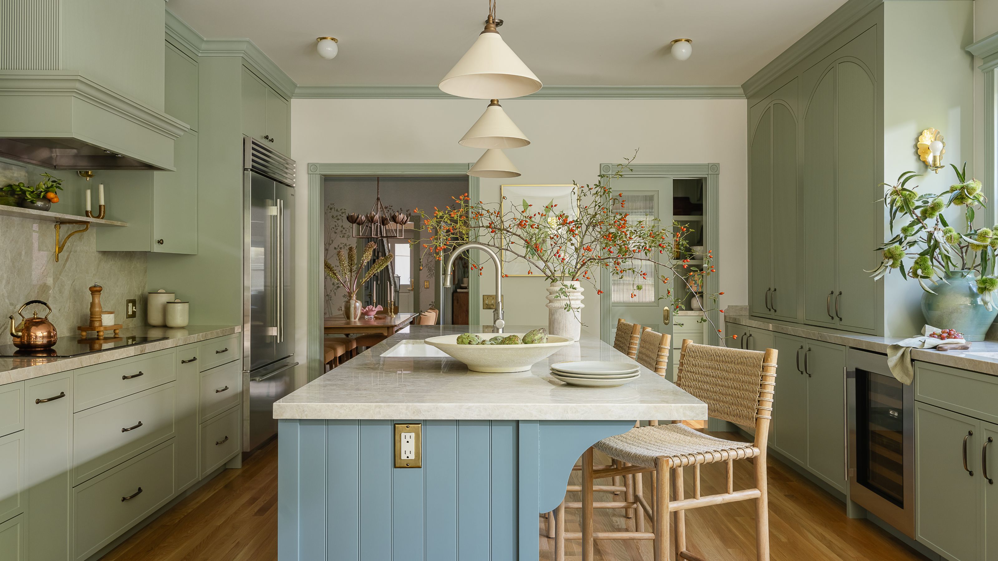 A kitchen with sage green cabinets, a large island with a blue paneled base and a light countertop, featuring woven bar stools and an open view to a dining room