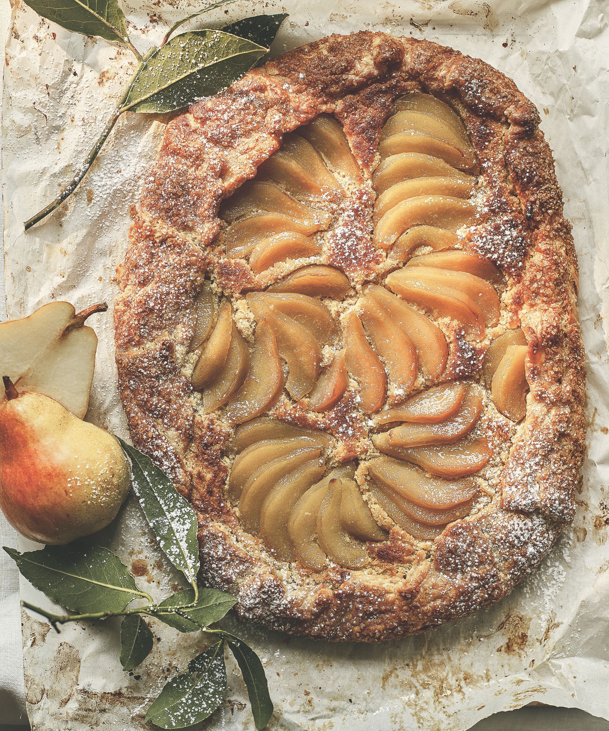 Pear tart on baking paper with whole pears and bay leaves to the side of the shot