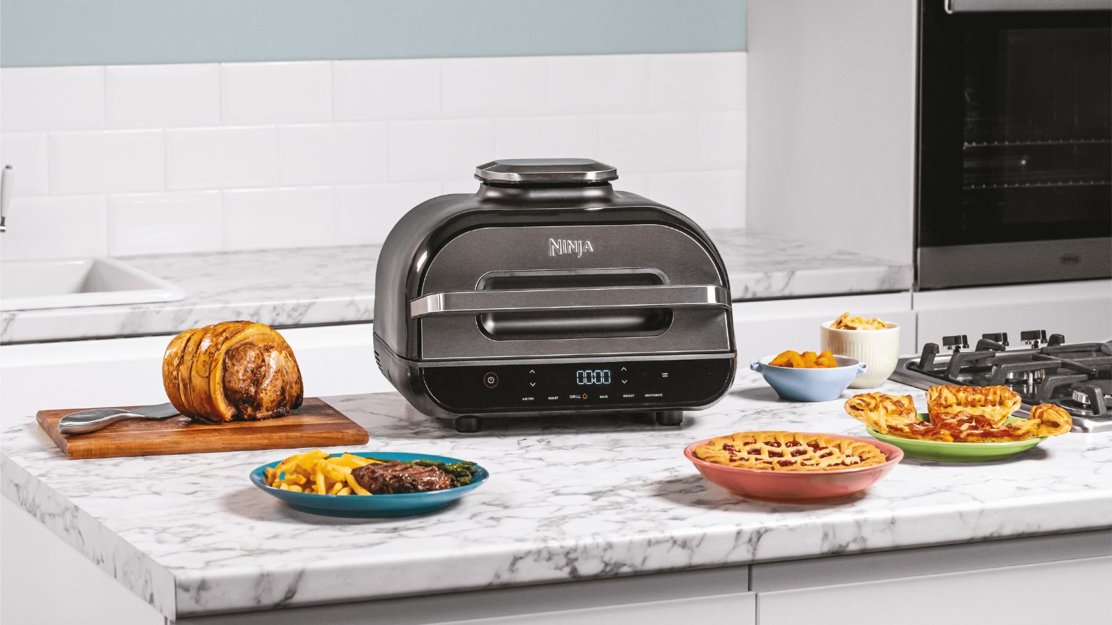 Ninja indoor grill on a kitchen counter surrounded by plates of food on a white marble worktop. 