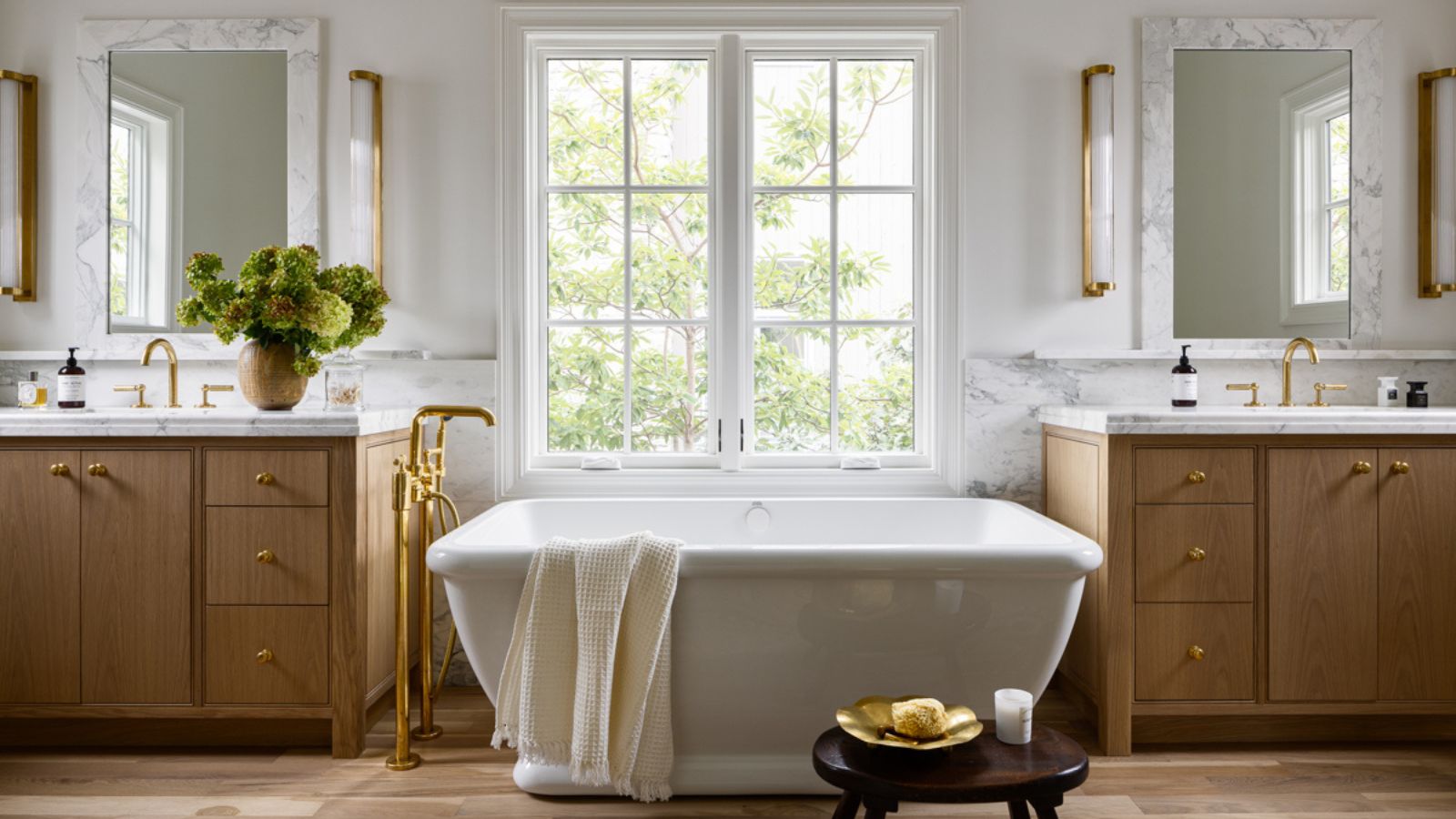 A large bathroom with walls painted Swiss Coffee by Benjamin Moore, two wooden vanities, and a freestanding bath beneath the window